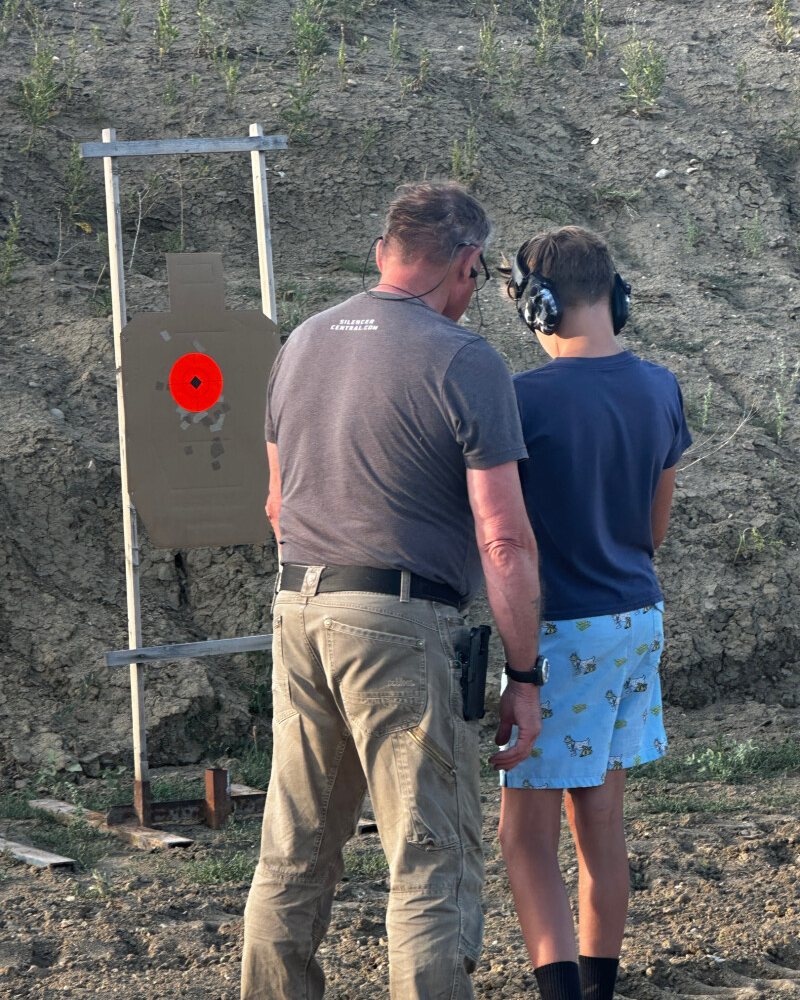 An ESS instructor coaching a boy wearing ear protection beside a silhouette target on a stand in front of the dirt berm