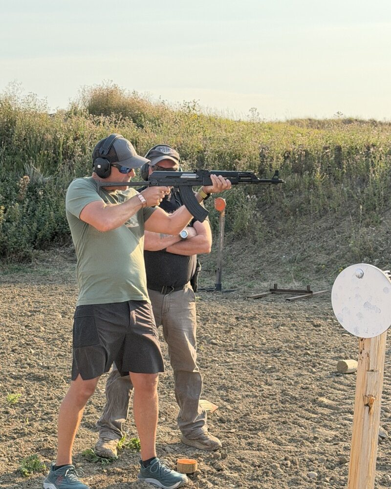 A shooter firing a rifle at steel targets in an ESS range bay while an instructor stands close behind him, both wearing ear protection, in evening light