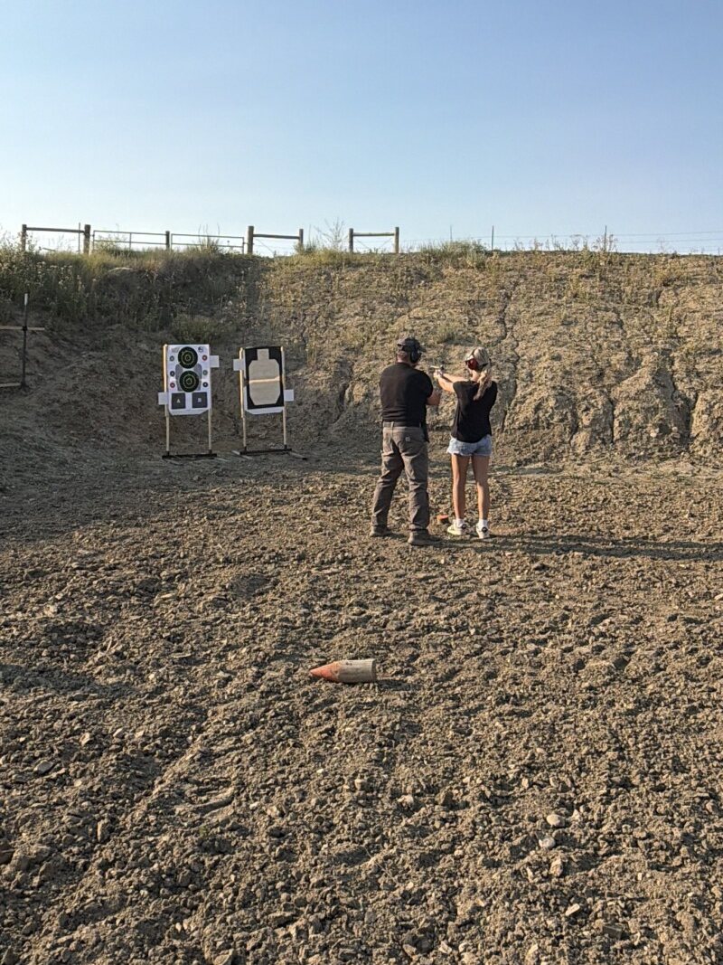 An instructor and a young woman shooting at paper targets in an open ESS bay below the big dirt berm, fence line and blue Montana sky above